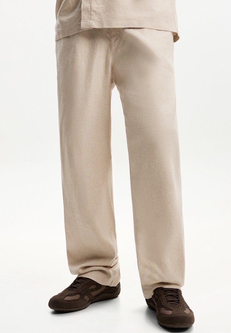 Pantalon ample beige porté avec des chaussures de ville décontractées marron foncé à lacets sur un fond blanc uni.