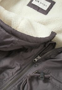 Šedá vnější látková bunda s jemným bílým fleecem uvnitř, obsahující přední zip, nastavitelné stahování a štítek.