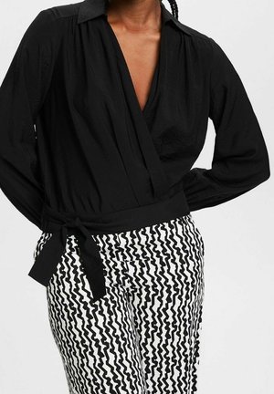 Vrouw die een zwarte lange mouw overslagblouse draagt, gestrikt in de taille, en zwart-witte zigzagprint broek.