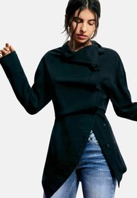 Cămașă neagră cu mâneci lungi, cu un tiv asimetric, realizată dintr-un material neted; are un guler înalt și închidere cu nasturi pe față.