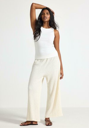 Vit ribbad linne topp kombinerad med lösa, texturerade off-white vida byxor och öppna sandaler med platt remdesign.