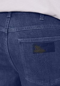 Tamno plave traperice s kožnom oznakom s logotipom "Wrangler", šavovima na stražnjim džepovima u obliku chevrona i suptilnom teksturom.