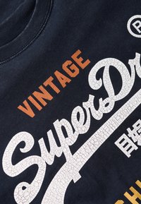 Marineblauw katoenen t-shirt met "VINTAGE" in oranje en "SuperDry" in gescheurd witte letters, met extra tekst in wit en gouden accenten.