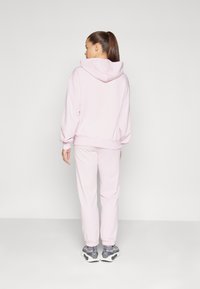 Lyserød hoodie med en baglomme og lange ærmer, parret med matchende joggers. Har elastiske manchetter og grå træningssko.