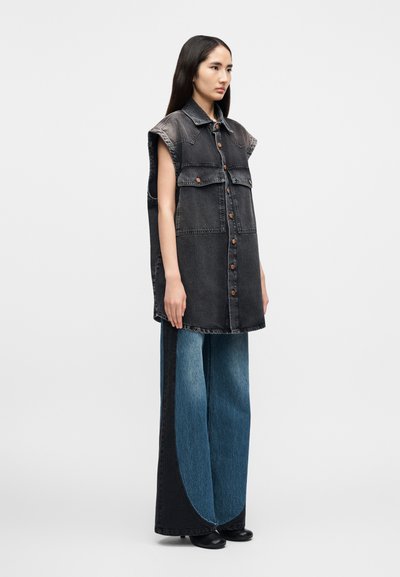 Ξεπερασμένο μαύρο γιλέκο από τζιν με κουμπιά και γιακά, με δύο μπροστινές τσέπες, συνδυασμένο με παντελόνια τζιν φαρδιάς γραμμής σε σκουρό και ανοιχτό μπλε χρώμα με διαβάθμιση.