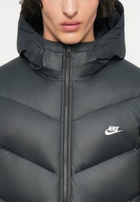 Chaqueta acolchada negra con capucha, con un diseño de acolchado en forma de chevrón, cremallera delantera y logo blanco de Nike en el lado izquierdo del pecho.