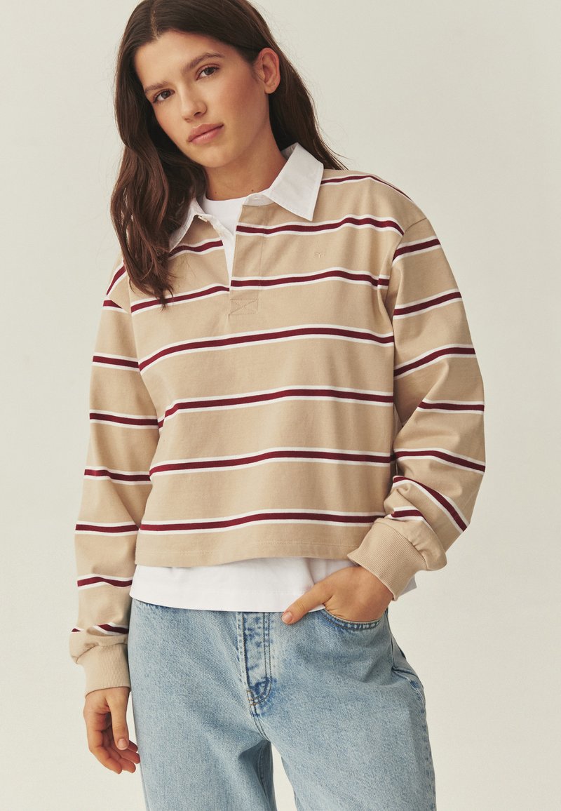Jeune femme aux longs cheveux bruns portant un maillot de rugby à rayures beige et bordeaux, un sous-maillot blanc et un jean bleu clair, main dans la poche.