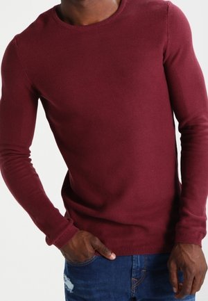 Homme portant un pull en maille ajusté bordeaux et un jean bleu déchiré, avec une main dans la poche sur un fond blanc uni.