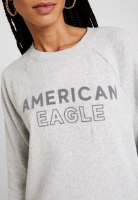 Felpa grigia con scollo rotondo e maniche raglan. Presenta una stampa audace "AMERICAN EAGLE" in un grigio scuro a contrasto. Tessuto morbido e testurizzato.