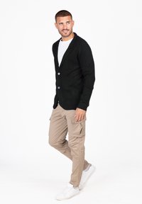 Cardigan nero con colletto a scialle, bottoni e tasca frontale, abbinato a pantaloni cargo beige e sneakers bianche. Tessuto morbido e design aderente.