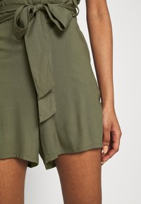 Olive green romper in lightweight fabric, featuring a tie belt and a loose fit. The design includes a layered panel on the front.