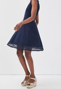 Robe sans manches bleu marine avec une jupe évasée et une bordure en dentelle à l'ourlet. Sandales compensées métalliques dorées avec texture tissée et détail de boucle.