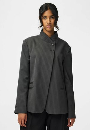 Femme aux cheveux foncés portant un blazer gris foncé à fines rayures avec un col asymétrique et deux boutons près du cou.
