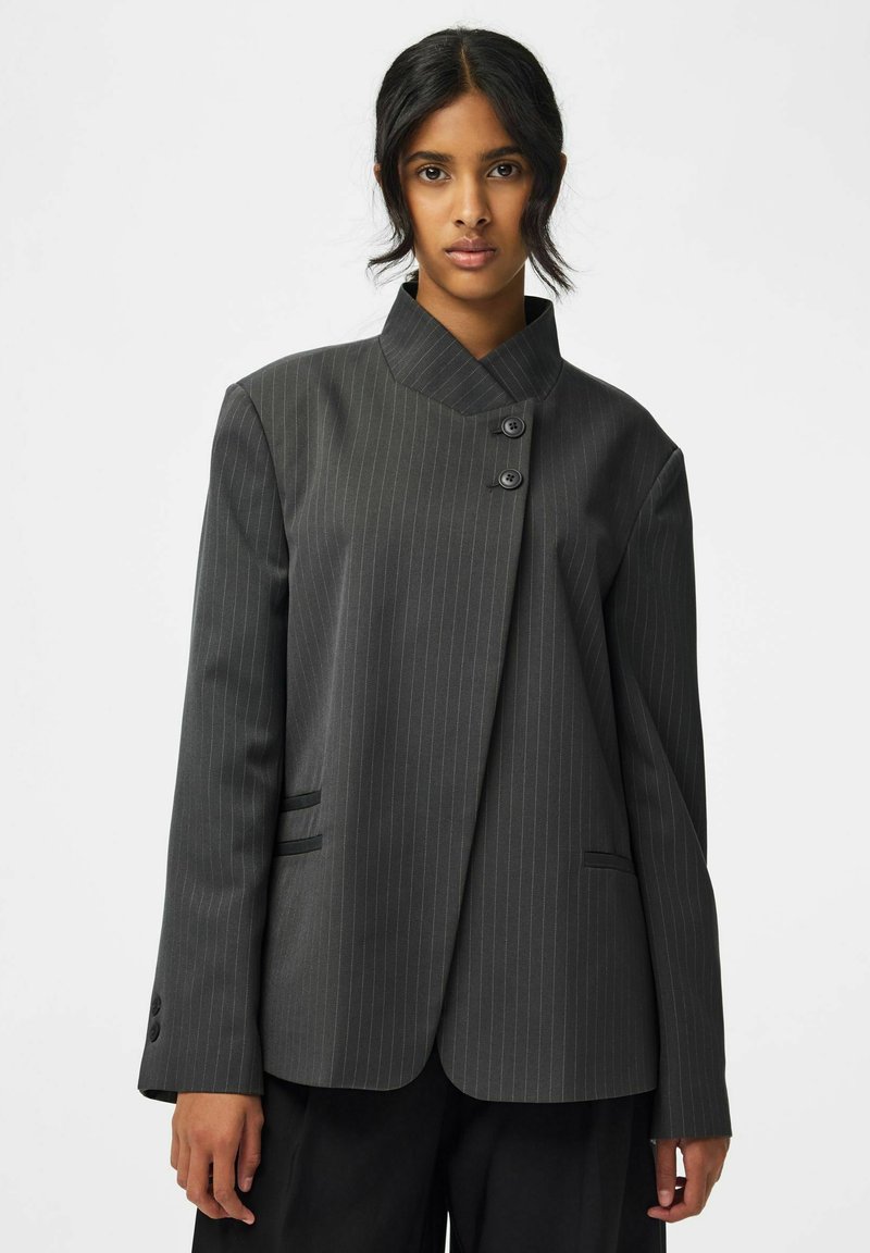 Femme aux cheveux foncés portant un blazer gris foncé à fines rayures avec un col asymétrique et deux boutons près du cou.