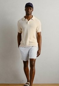 Beige knitted polo shirt with short sleeves, paired with textured light gray shorts. Model wears a navy cap and sandals.