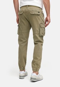 Uomo che indossa pantaloni cargo verde oliva con tasche a pattina e sneakers bianche New Balance, visto da dietro su sfondo bianco.