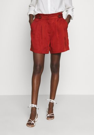 Woman wearing high-waisted red shorts with buttons and pleats, paired with a white tucked-in blouse and white strappy sandals.