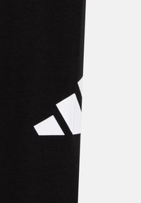 Un tissu noir avec un logo Adidas partiel en blanc, comportant trois bandes diagonales sur le côté droit, le tout sur un fond blanc.
