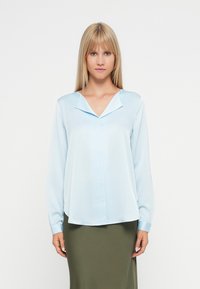 Svetlo modra bluzica z dolgimi rokavi in V-izrezom ter zloženim ovratnikom; gladka tekstura; v kombinaciji s prilegnjeno olivno zeleno krilo.
