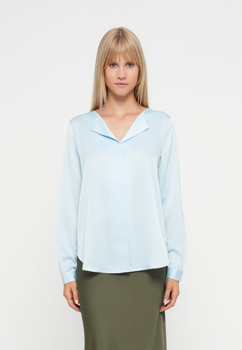 Svetlo modra bluzica z dolgimi rokavi in V-izrezom ter zloženim ovratnikom; gladka tekstura; v kombinaciji s prilegnjeno olivno zeleno krilo.