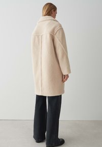 Cappotto in pelliccia sintetica di un morbido beige, design oversize con ampio colletto, spalle scese e dettagli sulle cuciture. Abbinato a pantaloni neri a gamba larga.