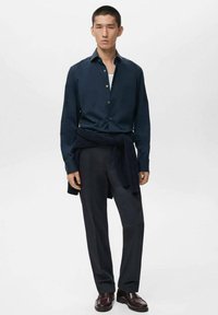 Camisa de botones azul marino en tejido suave, combinada con pantalones oscuros y zapatos burdeos. Suéter atado a la cintura, diseño minimalista.