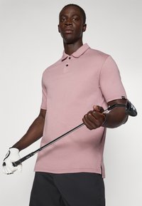 Polo rose clair, à manches courtes, avec un col et une patte de boutonnage à trois boutons. Le modèle tient un club de golf, portant un gant blanc.
