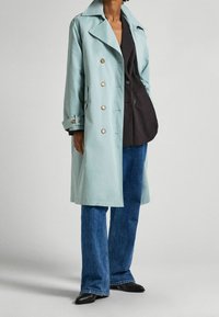 Jasnoniebieski wodoodporny prochowiec z szerokim kołnierzem, dwurzędowym zapięciem i złotymi guzikami, w zestawieniu z ciemnymi jeansami i czarnymi butami.