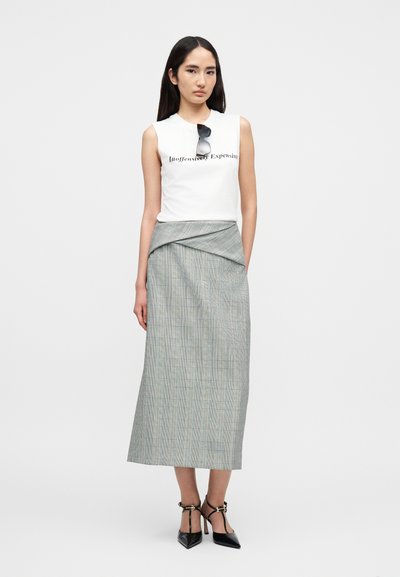 Λευκή αμάνικη μπλούζα με τυπωμένο κείμενο, συνδυασμένη με γκρι φούστα τύπου wrap με καρό σχέδιο και μαύρα πέδιλα με λουριά. Η φούστα έχει πλισέ λεπτομέρειες.