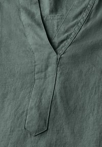Tessuto in lino verde chiaro con un design a V e un placket appuntito. La texture è leggermente ruvida e naturale, che mostra un aspetto informale.