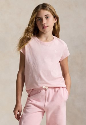 Jeune fille aux longs cheveux blonds portant un t-shirt rose clair à manches courtes et un pantalon de survêtement assorti sur un fond neutre.