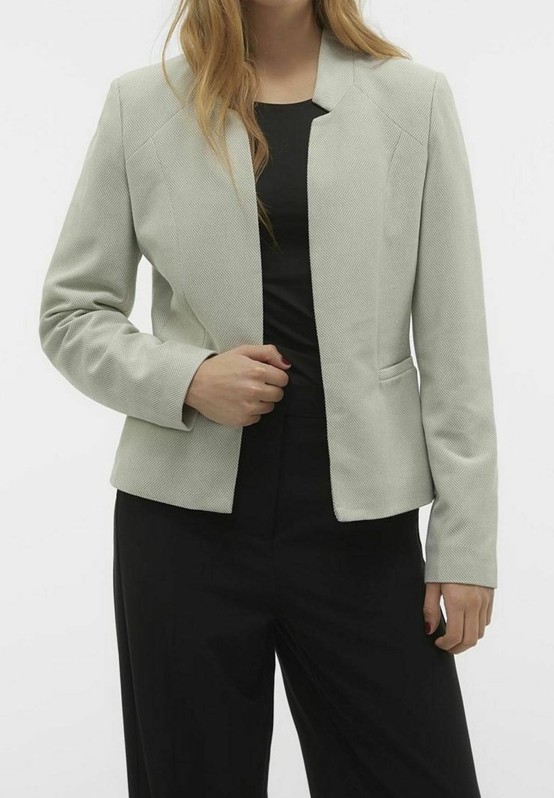 Femme portant un blazer gris clair texturé sur un haut noir et un pantalon noir, debout avec une main près de l'ouverture du blazer.