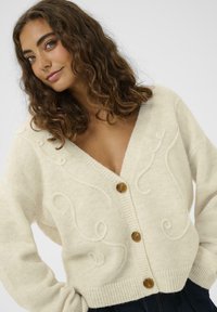 Krem boje kardigan s v-izrezom, rebrastim rubom i dekorativnom uvijenom vezom. Pet smeškastih dugmadi služi za zatvaranje.