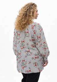 Blouse florale blanche avec des rayures verticales noires. Présente des fleurs roses et bleues, des manches bouffantes longues et une coupe décontractée. Tissu texturé.