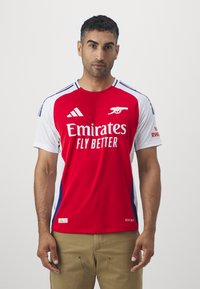 Maillot de football rouge et blanc avec manches courtes, portant le logo "Emirates Fly Better", des rayures Adidas et un matériau texturé.