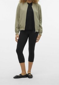 Oliwkowa kurtka bomberka, czarna bluzka, czarne legginsy 3/4 oraz czarne baleriny z ozdobą w postaci kokardy, stojąca na jednolitym tle.