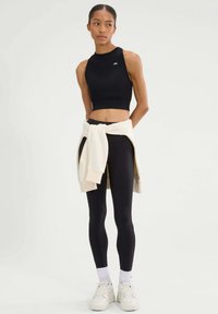 Zwarte cropped tanktop met een hoge halslijn, gecombineerd met zwarte leggings. Een crèmekleurige sweatshirt is om de taille gebonden. Witte sneakers maken de look compleet.
