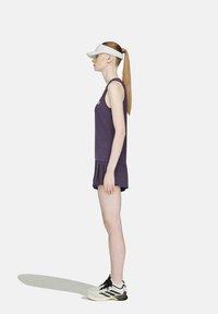 Frau mit langem Pferdeschwanz, die eine weiße Sonnenblende, ein lila ärmelloses Kleid und weiße Sneakers trägt, steht seitlich vor einem einfarbigen Hintergrund.