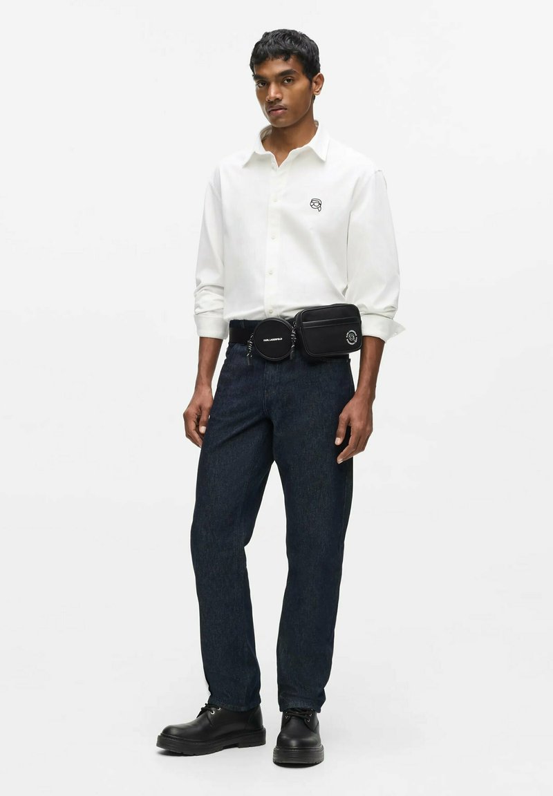 Homme portant une chemise blanche boutonnée, un jean foncé, un sac banane noir à deux compartiments et des chaussures noires à lacets, debout devant un fond blanc.