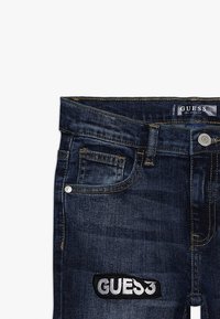 Tmavě modré džíny s přiléhavým střihem, s vyšitým štítkem s nápisem "GUESS" v bílé barvě na černé nášivce a stříbrnými doplňky v oblasti pasu.