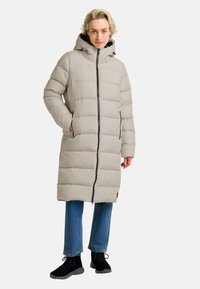 Licht beige pufferjas met een capuchon, voorzien van een voorkant met rits, zijzakken en een knielange snit. Draag het over een blauwe spijkerbroek en zwarte schoenen.