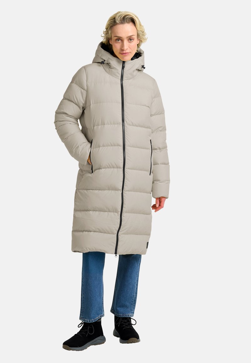 Licht beige pufferjas met een capuchon, voorzien van een voorkant met rits, zijzakken en een knielange snit. Draag het over een blauwe spijkerbroek en zwarte schoenen.