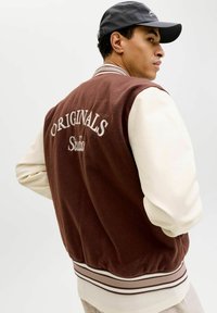 Brun varsityjakke i ullblanding med kremfargede ermer, med «ORIGINALS» og «Studio» brodert på baksiden. Ribbestrikkede mansjetter og kant.