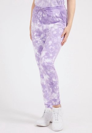 Femme portant un pantalon de survêtement tie-dye violet et blanc avec taille et poignets élastiques, associé à des baskets blanches, debout devant un fond uni.