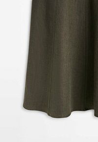 Jupe en tissu vert olive foncé avec coutures visibles suspendue devant un fond blanc.