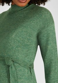 Maglione verde lavorato a maglia con collo alto, maniche lunghe e cintura in tessuto. La trama appare morbida e leggermente melangiata.