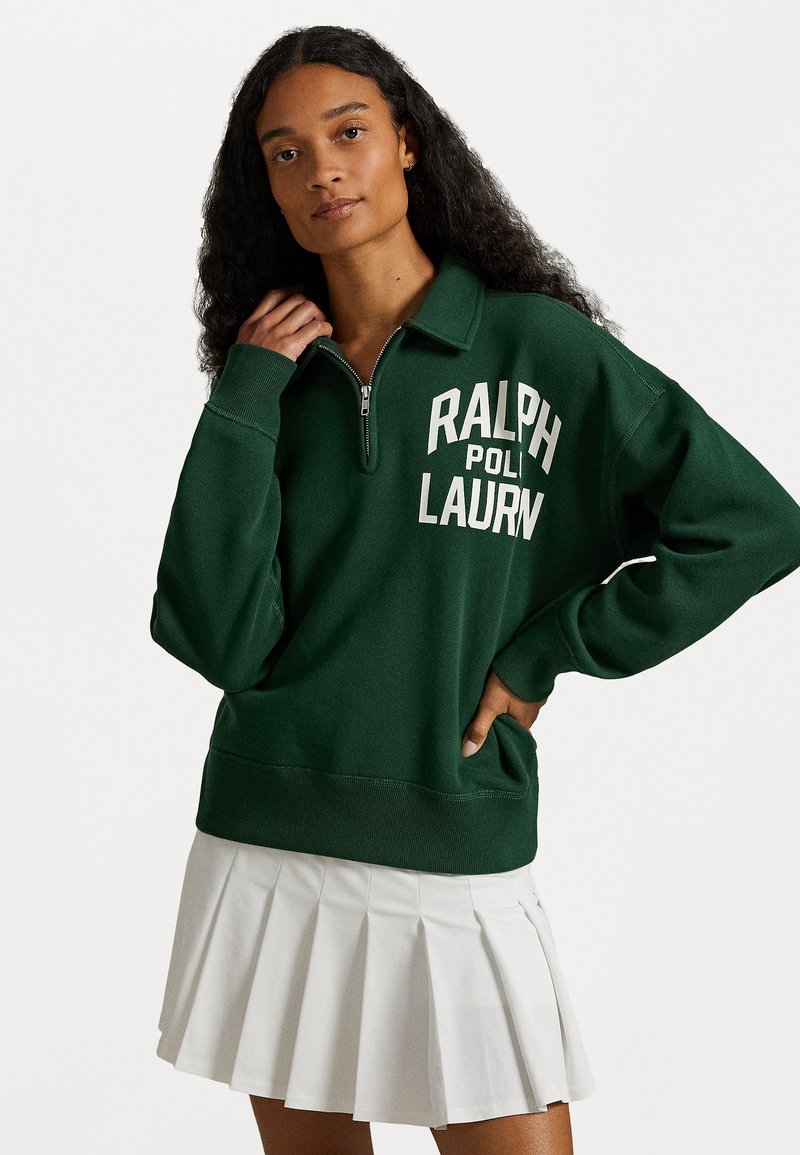 Green sweatshirt with a quarter-zip collar and white "RALPH LAUREN" text. Soft, textured fabric with a relaxed fit, paired with a pleated white skirt.