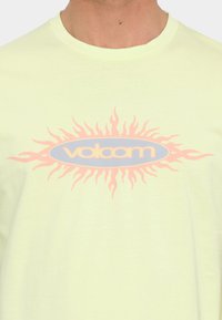 Camiseta de algodón amarillo claro con un gráfico central de llamas en rosa y azul con la palabra "Volcom" en el centro. Mangas cortas, cuello redondo.