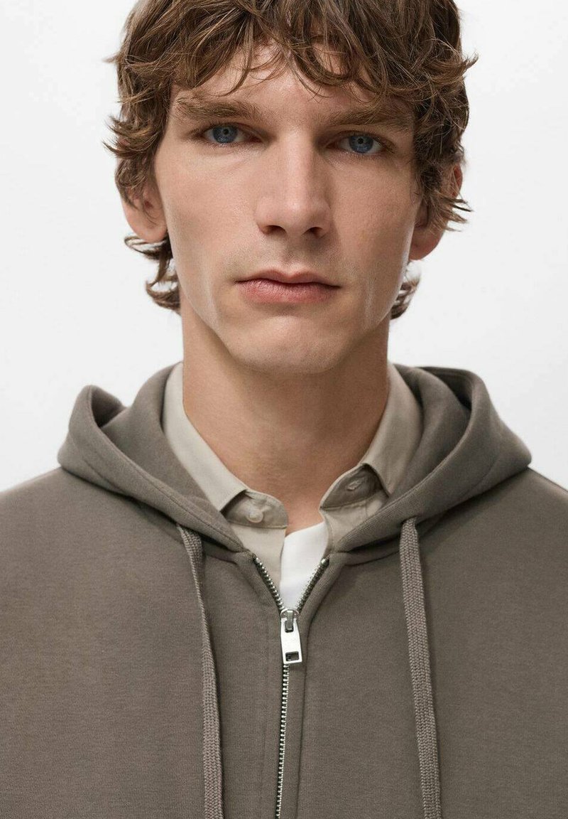 Brown zippered hoodie with a drawstring, layered over a light shirt. The fabric appears soft with a smooth texture and neutral tones.