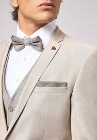 Veste beige en tissu lisse, associée à un nœud papillon à motifs gris clair et à un gilet gris, avec un mouchoir de poche et une épingle à revers.